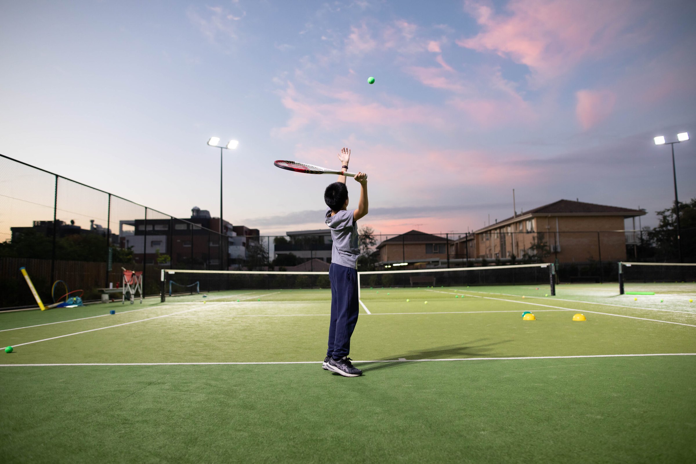 A junior FD Tennis player serving at sunset on the synthetic grass courts